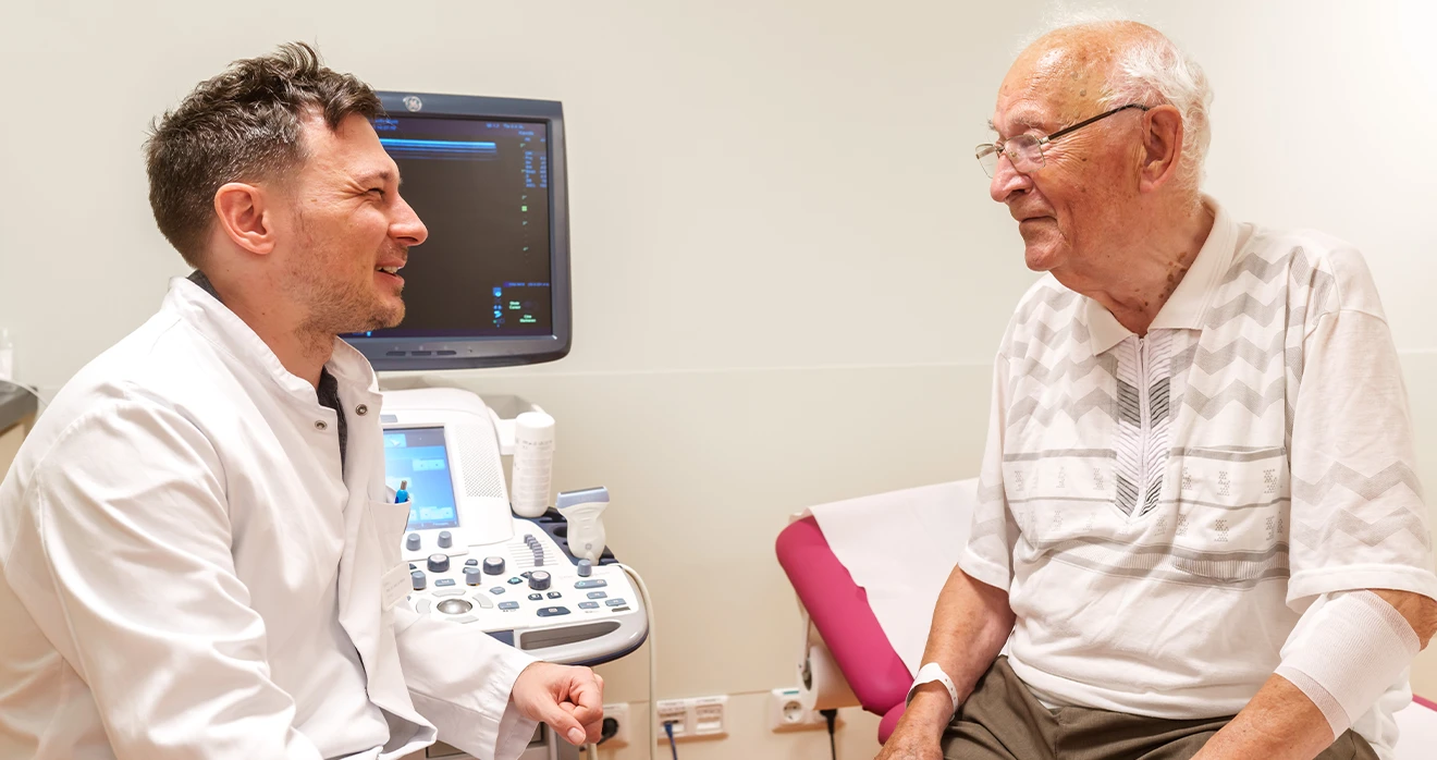 Ein Arzt lächelt einem älteren Patienten zu, der gerade auf einer Liege in einem Behandlungsraum sitzt.