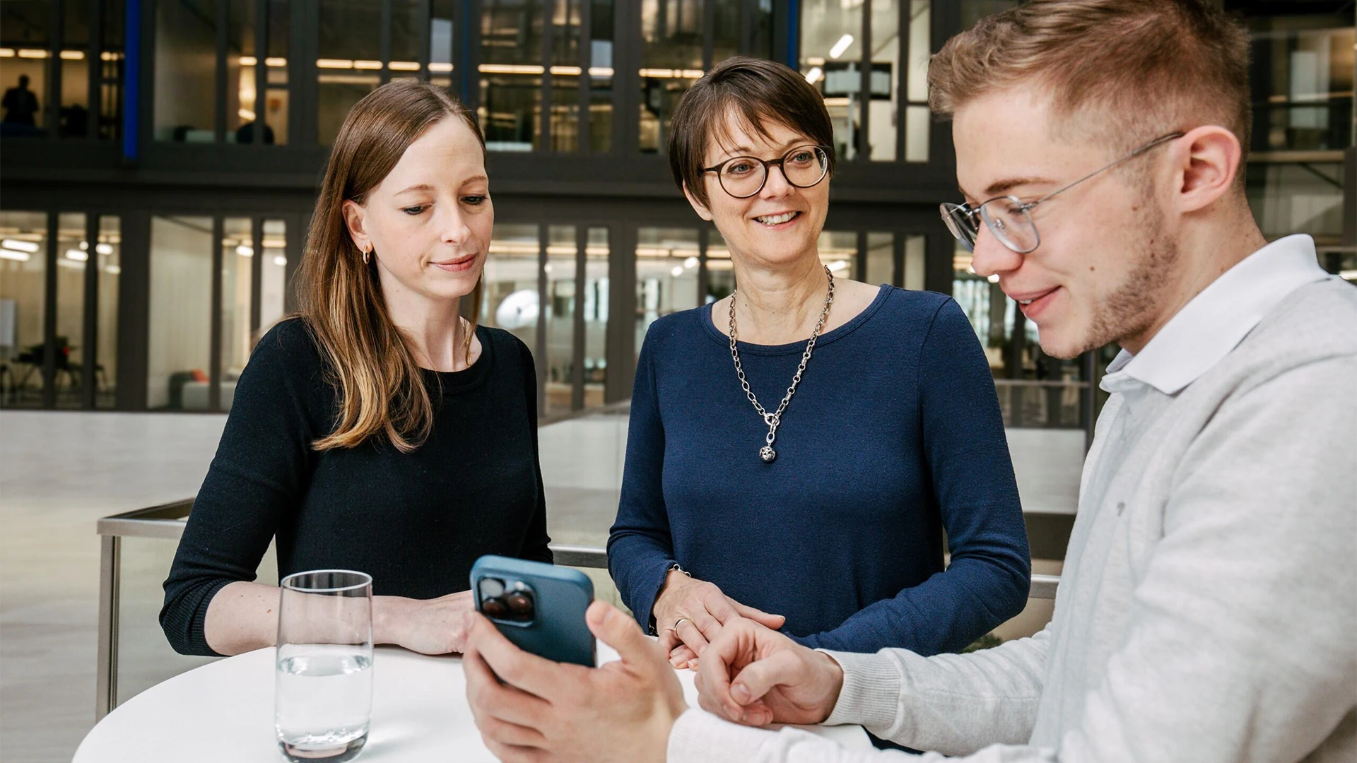 Drei personen stehen um eine Stehtisch. Ein Mann hält ein Handy in Richtung der anderen beiden. Die eine Frau sieht auf dieses Handy, während die andere den Mann anschaut.