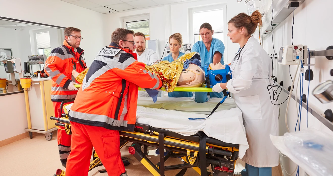 Sanitäter platzieren eine dummy Puppe auf eine Trage, während medizinisches Personal um sie herumsteht. Es wirkt wie eine Fort- und Weiterbildung oder ein Training.