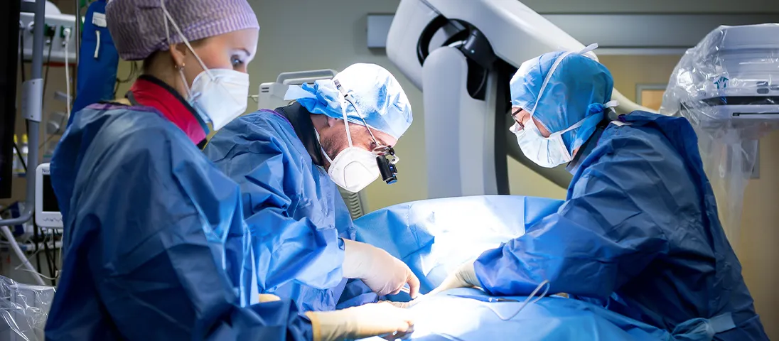 A surgical procedure appears to be taking place where two surgeons are performing an operation, with one standing at the sterile field and the other working at a surgical equipment station.