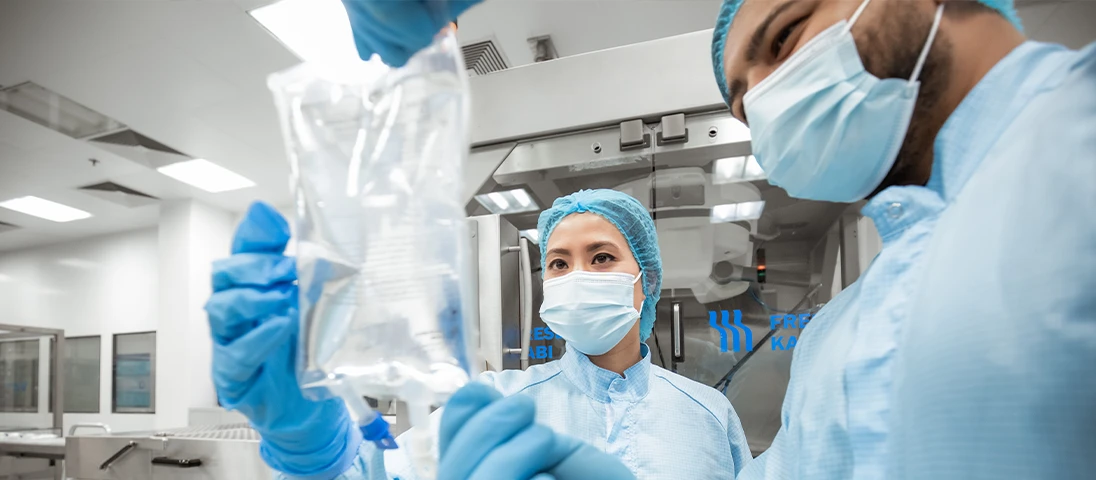 A healthcare factory worker in protective gear is holding a transparent infusion bag. Another individual, also in medical attire, is standing nearby, looking at the contents of this bag with interest