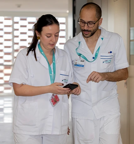 Two individuals, likely nurses or healthcare assistants. They are both standing and attentively looking at a phone with the screen facing them, possibly communicating or viewing some information relevant to their work.
