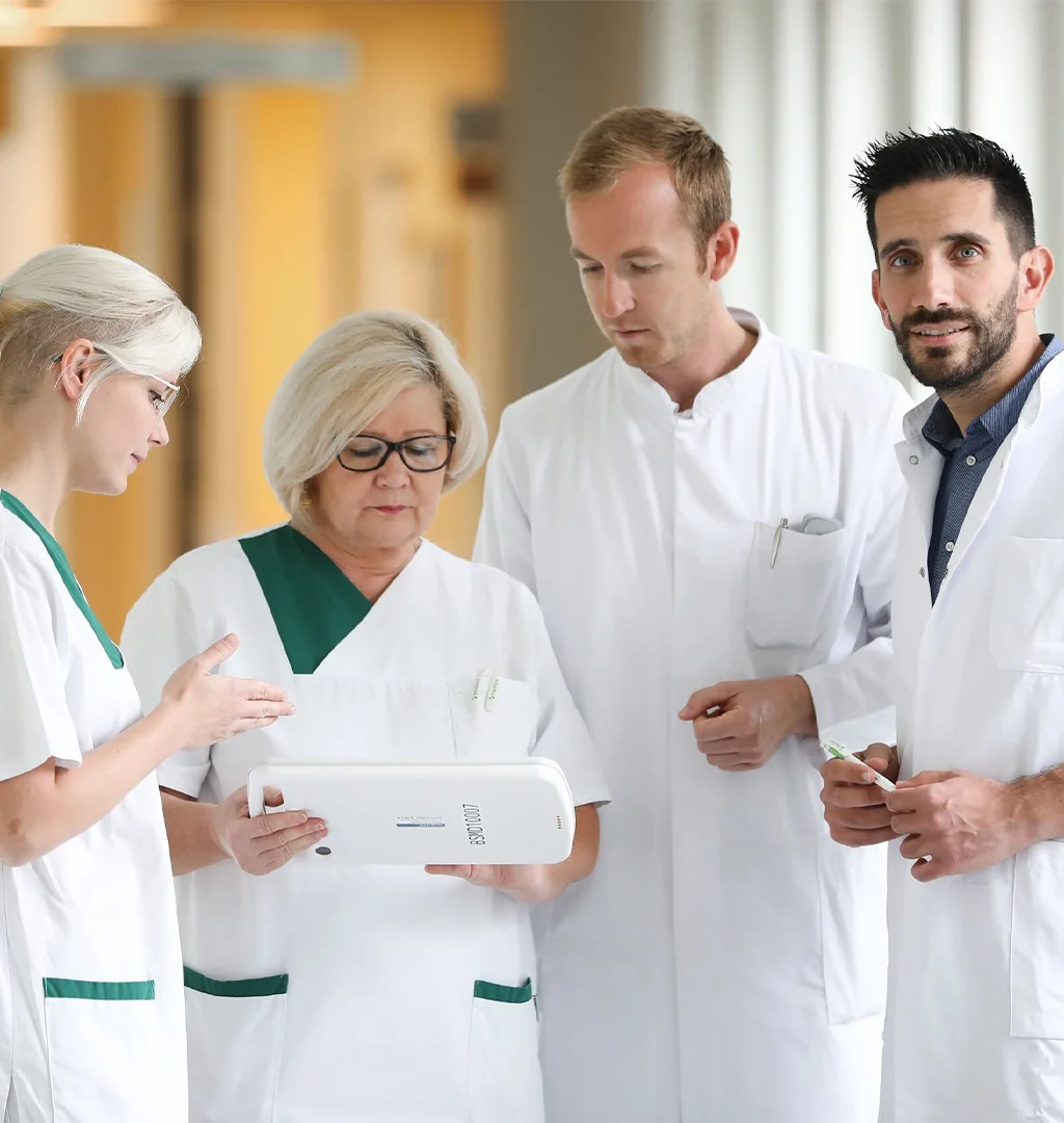 The image shows a group of healthcare workers or professionals in what appears to be a hospital setting. One person is holding a tablet, all the individuals seem focused on what they are doing or discussing.The person on the right is looking straight into the camera.
