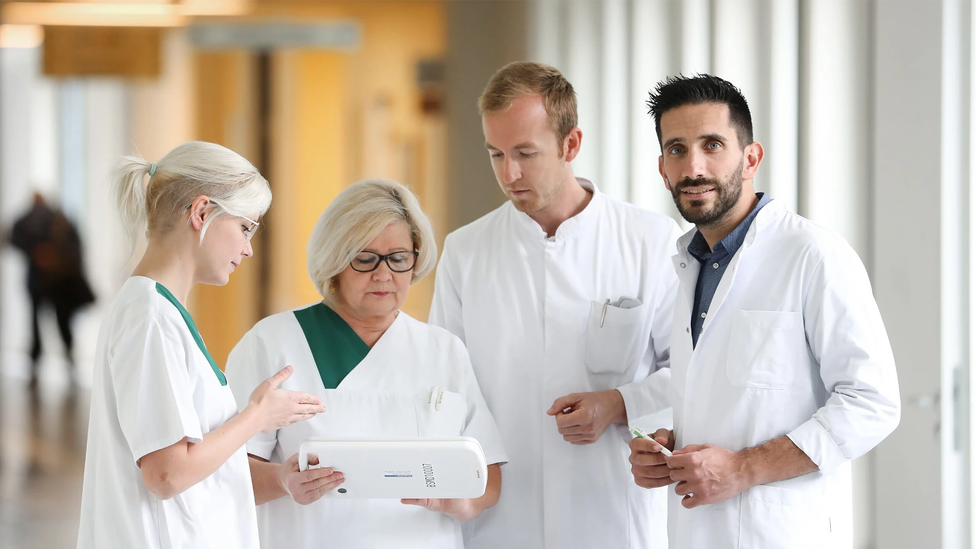 The image shows a group of healthcare workers or professionals in what appears to be a hospital setting. One person is holding a tablet, all the individuals seem focused on what they are doing or discussing.The person on the right is looking straight into the camera.