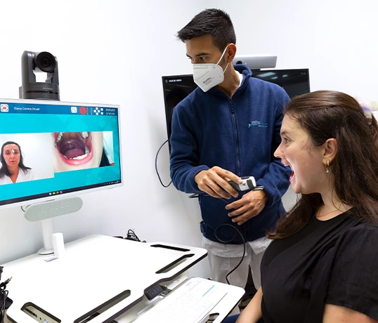 A healthcare worker films the mouth of a patient. The filming appears on a screen in front of them, which also shows another healthcare worker participating in a video call.