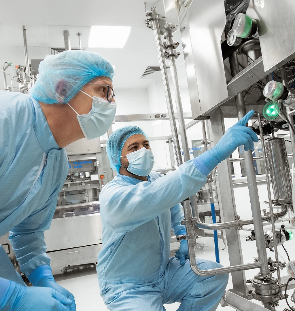 In the image, there are two individuals wearing blue lab coats and hair nets. They are working with industrial-scale equipment. The person on the left is leaning towards the machine to either operate it or inspect its contents, while the other individual stands to the side pointing at something.