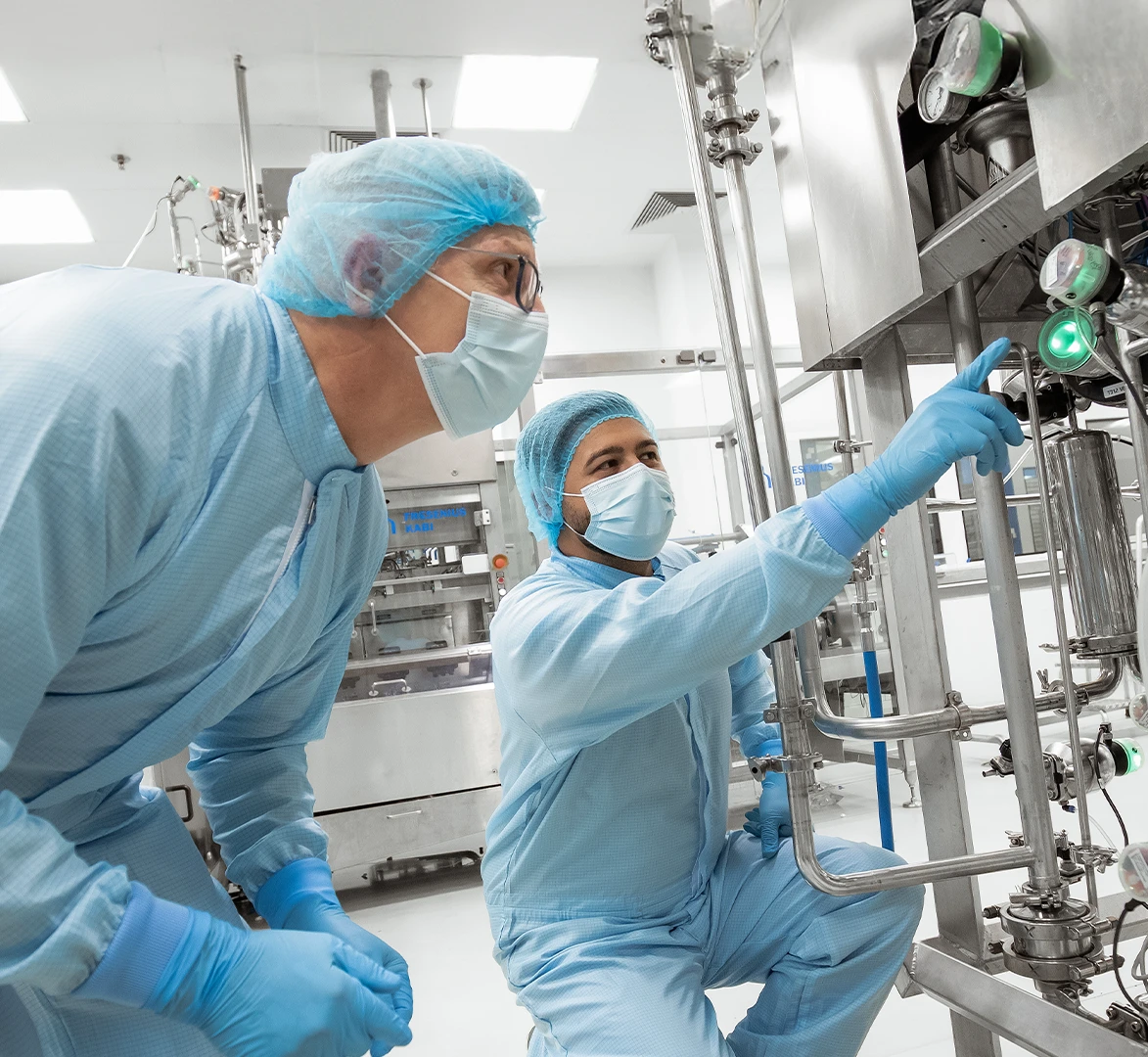 In the image, there are two individuals wearing blue lab coats and hair nets. They are working with industrial-scale equipment. The person on the left is leaning towards the machine to either operate it or inspect its contents, while the other individual stands to the side pointing at something.