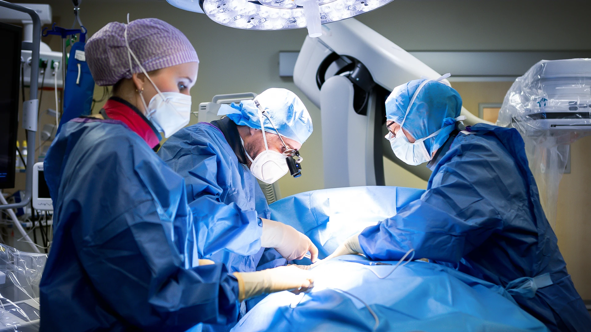 The image shows healthcare professionals engaged in a medical procedure. They are wearing surgical gowns, masks, and head caps for sterilization purposes to prevent the spread of bacteria during surgery.