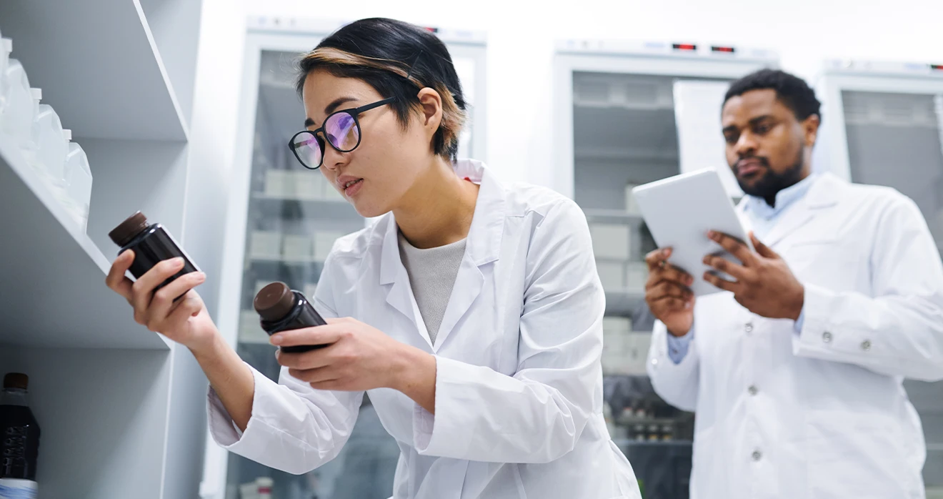 The image depicts two individuals in what appears to be a laboratory setting. One person is holding two bottles. The other is looking at a tablet. They are wearing laboratory attire and glasses.
