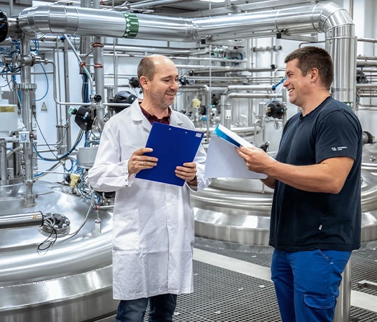 The image shows two individuals engaging in a discussion beside machinery.
