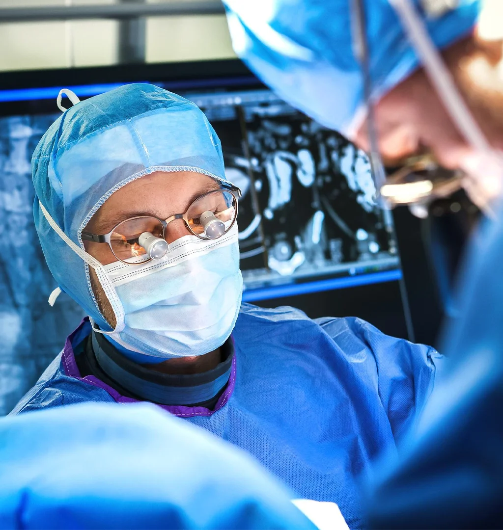 The image shows a person wearing surgical gown, cap, and goggles in an operating room environment.