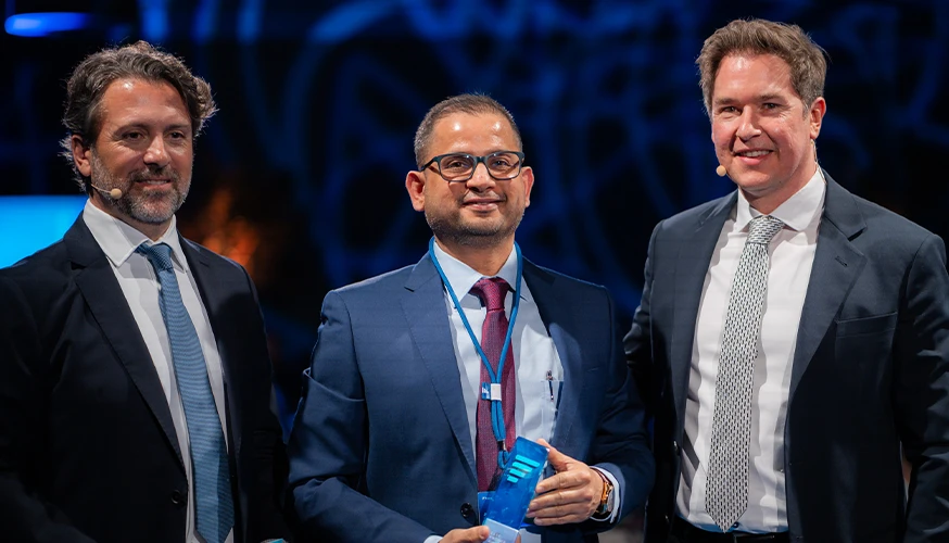 In the image, you can see three men standing side by side on a stage. The man in the center has an award plaque in front of him.