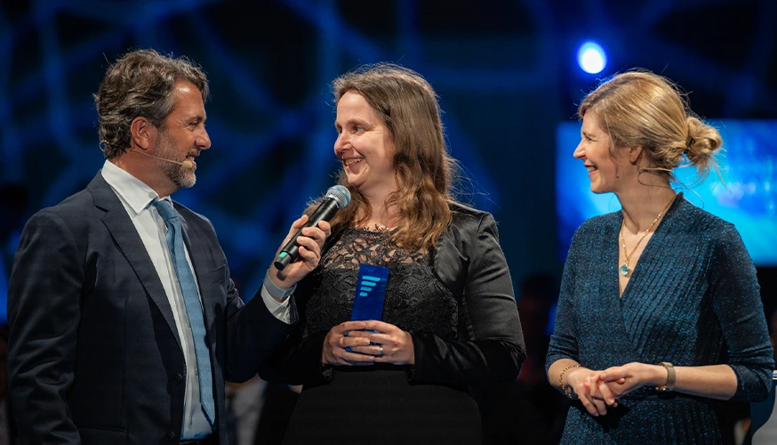 This is an image of three individuals. The woman in the center is holding an award and the man on the left is holding a microphone to the woman with the award. The woman on the right is smiling beside them.