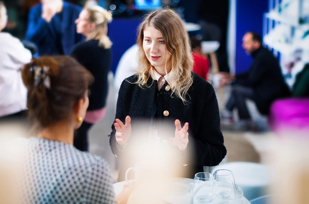 This image appears to be a photograph capturing a moment during an event. In the foreground, there is a person talking to another individual who has their back to the camera.