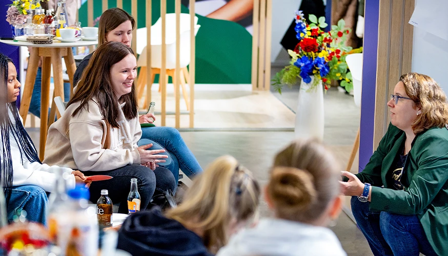This appears to be a photo of a group of people gathered. There is a person kneeling at the forefront of the image, interacting with others. In the background, you can see exhibition booths with graphics and displays on top of tables.