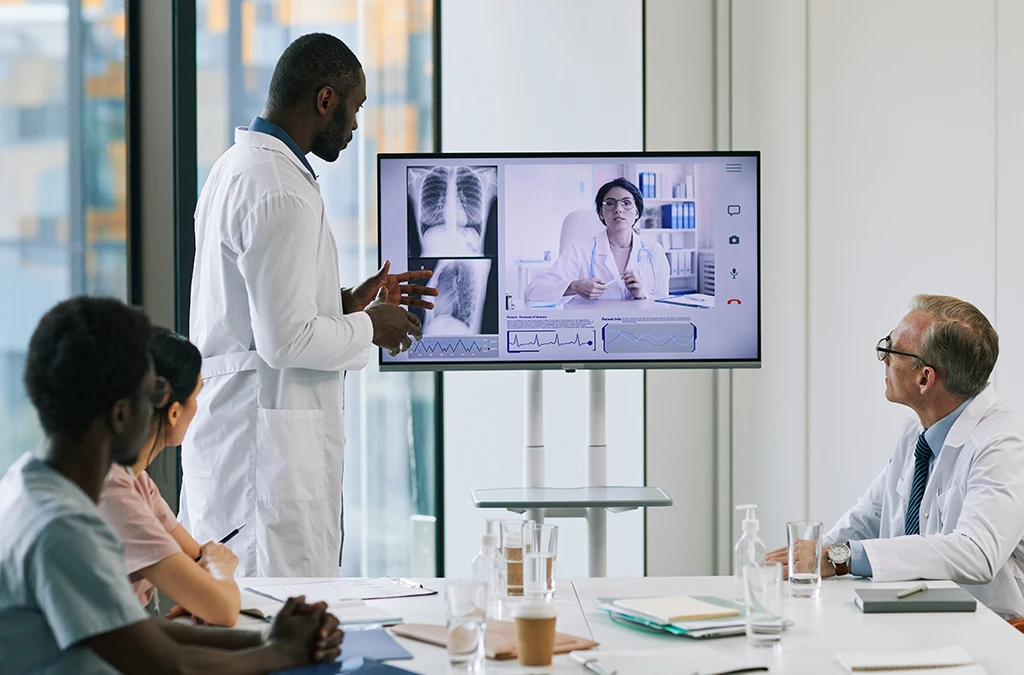 In the image, there is a scene where group of people are seated around a large screen displaying an X-ray image of a chest. The person at the front seems to be demonstrating something on the screen. The participants appear engaged in discussion or review.