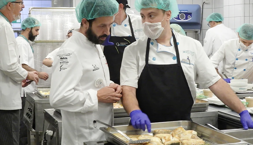 This image appears to be taken inside a kitchen or cafeteria where several cooks are preparing food. It shows cooks cooking and plating various kinds of food.