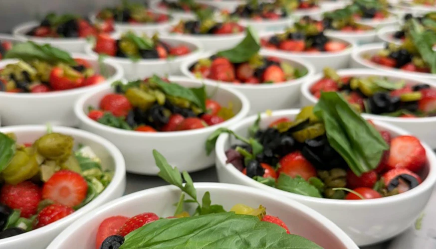 The image shows a tray of individual servings filled with fresh fruits such as strawberries and greens.