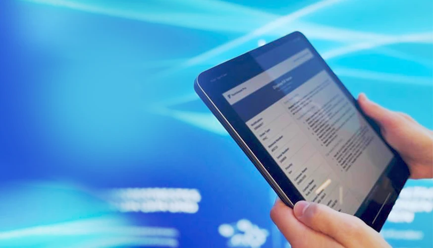 The image shows the screen of a tablet held with two hands. The person using it seems to be browsing information on the screen, which displays what appears to be a document or report with text.